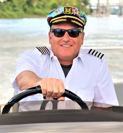 Smiling boat captain in a floral captain’s hat and sunglasses steering a motorboat on a sunny river waterway