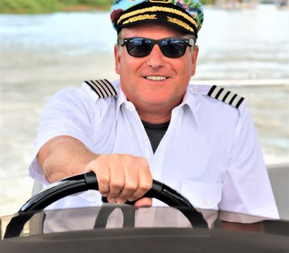 Smiling boat captain in a floral captain’s hat and sunglasses steering a motorboat on a sunny river waterway