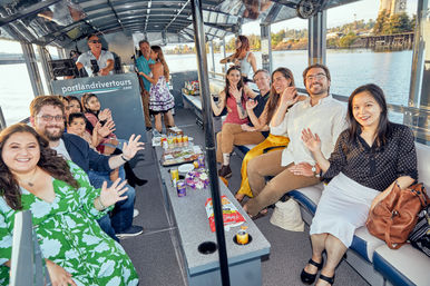 Smiling group waving aboard a covered sightseeing river cruise boat with snacks and drinks on the table and a sunlit waterfront city view.