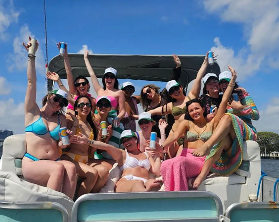 Group of friends in colorful bikinis and caps enjoying a sunny pontoon boat party, waving and holding canned drinks on blue coastal water