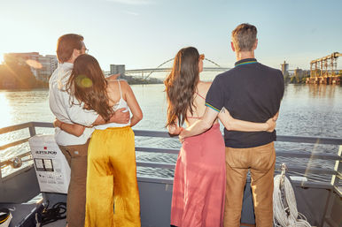 Two couples embracing on a boat deck, gazing across a sunlit urban river toward a steel arch bridge and city skyline at sunset.