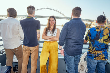 Sunlit river cruise scene: smiling woman in a white top and yellow wide-leg pants stands with arms crossed on a boat deck, flanked by four men facing the water and an arched bridge spanning the river in the background.