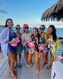 Six friends on a coastal docked boat by turquoise ocean at sunset celebrating a birthday, wearing casual summer outfits, birthday sash and tiara, holding drinks in pink flamingo float holders under a thatched tiki roof, one person holding a small white dog.