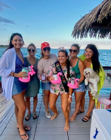 Six friends on a coastal docked boat by turquoise ocean at sunset celebrating a birthday, wearing casual summer outfits, birthday sash and tiara, holding drinks in pink flamingo float holders under a thatched tiki roof, one person holding a small white dog.