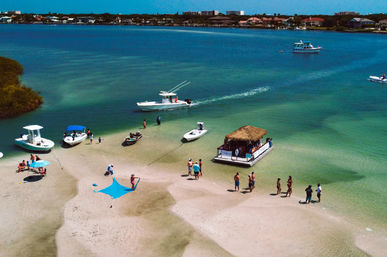 Aerial view of a sunny coastal sandbar with anchored boats, a thatched-roof tiki float, and people wading in clear turquoise water.
