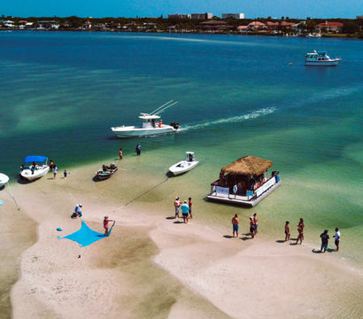 Aerial view of a sunny coastal sandbar with anchored boats, a thatched-roof tiki float, and people wading in clear turquoise water.