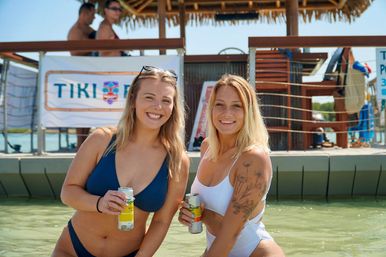 Two smiling women in swimwear holding canned drinks in shallow turquoise water by a tiki-style dock bar on a sunny beach day, vacation vibe
