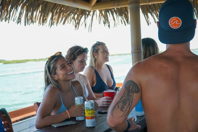 Smiling group at a tropical tiki-style bar on a boat, women in swimsuits holding canned drinks and a red cup, shirtless man with jellyfish tattoo and ocean in the background