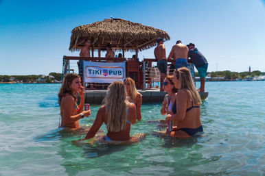 People in swimsuits wading in clear turquoise water around a floating tiki-style bar with a thatched roof, holding canned drinks on a sunny coastal day.