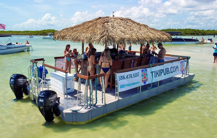 Floating tiki-bar pontoon with thatched roof and twin outboard motors anchored in shallow turquoise Florida waters by a sandbar, people in swimsuits socializing as nearby boats idle under a sunny sky.
