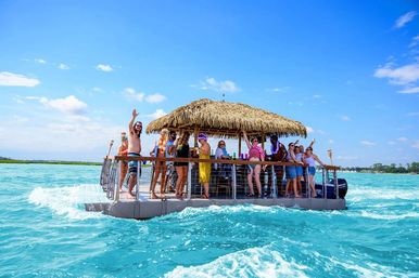 Cheerful group partying on a floating tiki-bar pontoon with a thatched roof in turquoise tropical waters under a bright blue sky