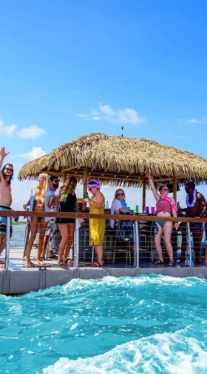 Cheerful group partying on a floating tiki-bar pontoon with a thatched roof in turquoise tropical waters under a bright blue sky