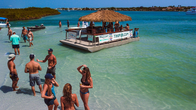 Sunny tropical sandbar with beachgoers in swimsuits socializing in clear turquoise water around an anchored thatched-roof floating tiki bar.