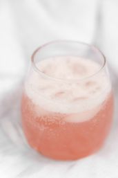 Close-up of a bubbly pink beverage with frothy foam in a stemless glass on a soft white fabric background.