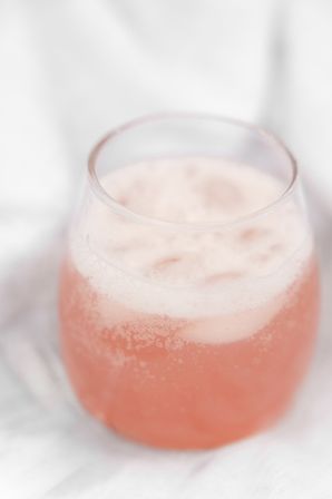 Close-up of a bubbly pink beverage with frothy foam in a stemless glass on a soft white fabric background.