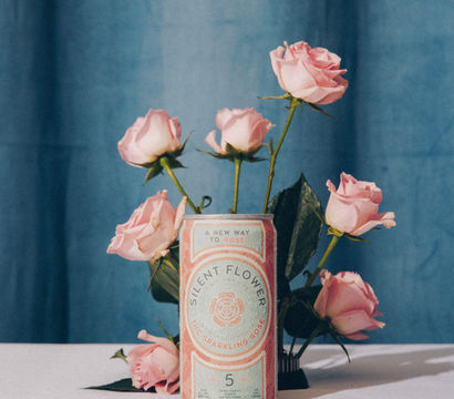 Pink roses arranged in a decorative can used as a vase on a white table, set against a soft blue curtain backdrop — romantic still-life, vintage aesthetic.