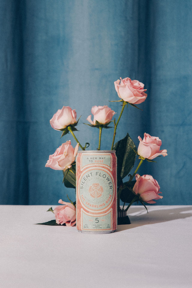 Pink roses arranged in a decorative can used as a vase on a white table, set against a soft blue curtain backdrop — romantic still-life, vintage aesthetic.