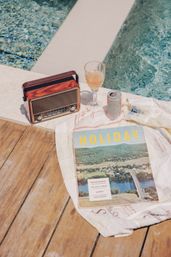 Sunlit poolside still life on a wooden deck featuring a vintage radio, a cocktail glass and can, and a 'HOLIDAY' travel magazine on a white towel beside a turquoise pool — dreamy summer escape.