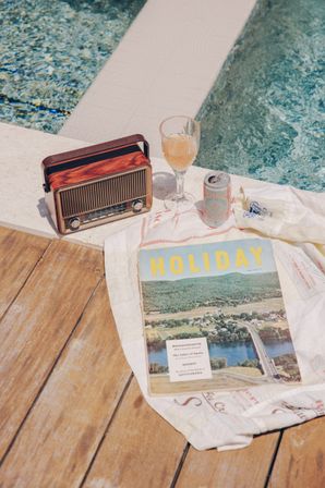 Sunlit poolside still life on a wooden deck featuring a vintage radio, a cocktail glass and can, and a 'HOLIDAY' travel magazine on a white towel beside a turquoise pool — dreamy summer escape.