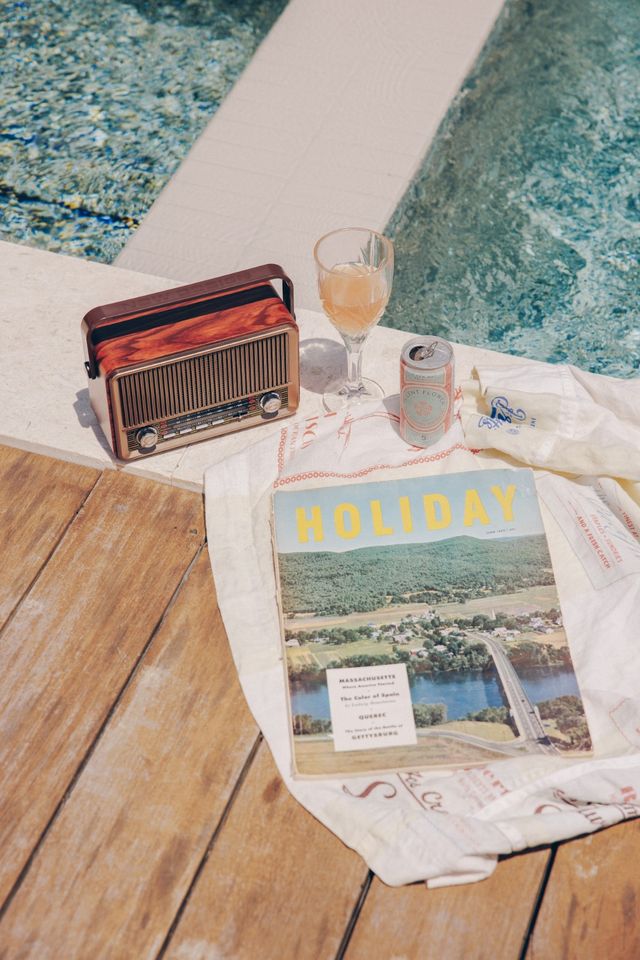 Sunlit poolside still life on a wooden deck featuring a vintage radio, a cocktail glass and can, and a 'HOLIDAY' travel magazine on a white towel beside a turquoise pool — dreamy summer escape.