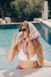 Woman in white bikini with striped headscarf and sunglasses sipping a canned drink while relaxing in a sunlit outdoor pool, summer poolside vibe