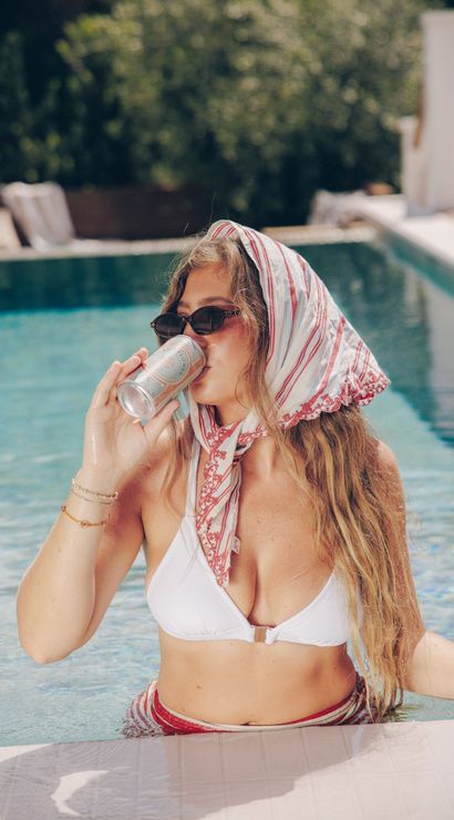 Woman in white bikini with striped headscarf and sunglasses sipping a canned drink while relaxing in a sunlit outdoor pool, summer poolside vibe