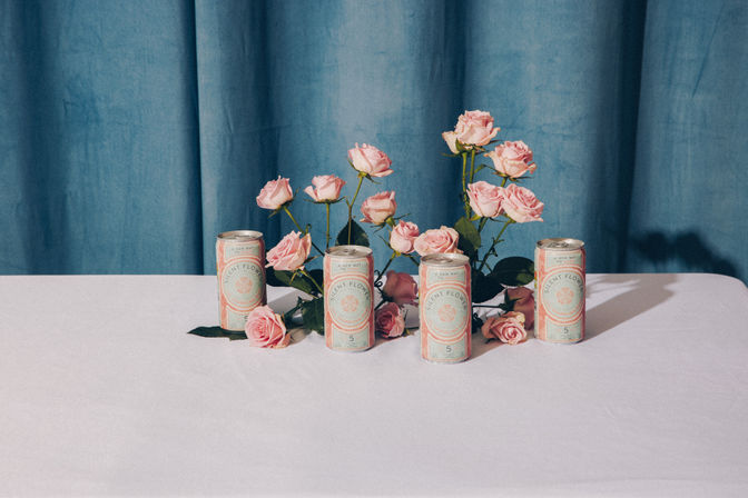 Chic still life of pink roses arranged with pastel pink-and-mint drink cans on a white table against a teal curtain backdrop