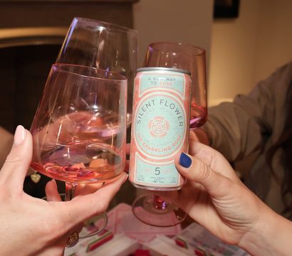 Close-up of three hands toasting pink wine glasses and a canned sparkling rosé beverage in a cozy living-room setting over a colorful tablecloth.