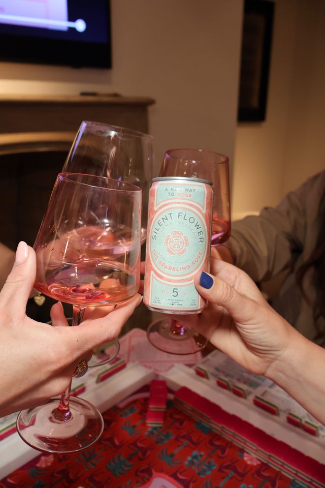 Close-up of three hands toasting pink wine glasses and a canned sparkling rosé beverage in a cozy living-room setting over a colorful tablecloth.
