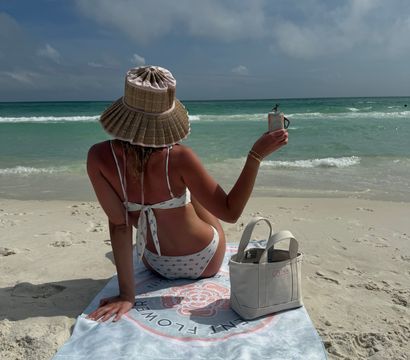 Person in a straw sun hat and white floral bikini sitting on a beach towel, raising a canned drink toward the turquoise ocean and partly cloudy sky, with a canvas tote on the white sand nearby.