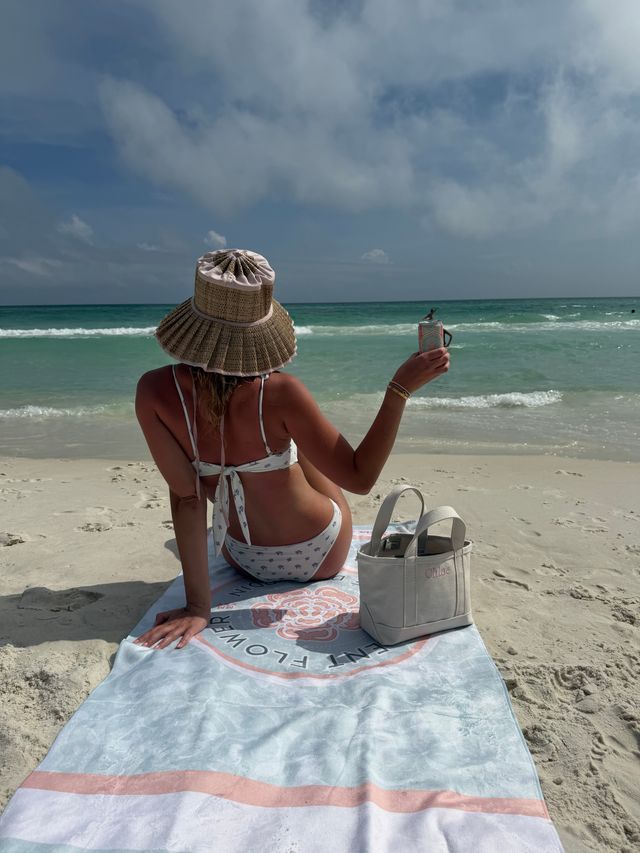 Person in a straw sun hat and white floral bikini sitting on a beach towel, raising a canned drink toward the turquoise ocean and partly cloudy sky, with a canvas tote on the white sand nearby.