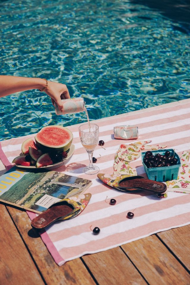 Poolside summer picnic on a wooden deck: pink-and-white striped towel with sliced watermelon, a carton of cherries, floral slide sandals and a glass being filled from a can beside sparkling blue pool water.