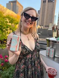 Smiling person in cat‑eye sunglasses and a floral dress on an urban rooftop terrace, holding a can of sparkling rosé and a cup with city skyline at sunset