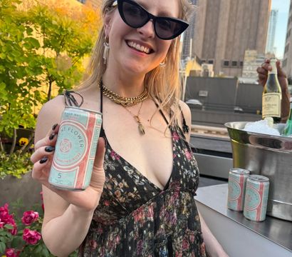 Smiling person in cat‑eye sunglasses and a floral dress on an urban rooftop terrace, holding a can of sparkling rosé and a cup with city skyline at sunset