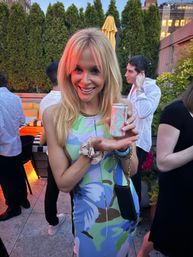 Smiling woman in a colorful floral dress holding a small canned drink on a city rooftop patio at dusk, with guests and tall greenery in the background.