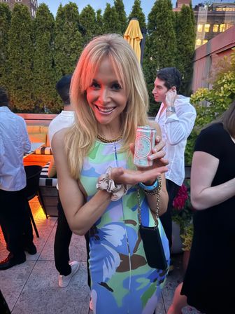 Smiling woman in a colorful floral dress holding a small canned drink on a city rooftop patio at dusk, with guests and tall greenery in the background.