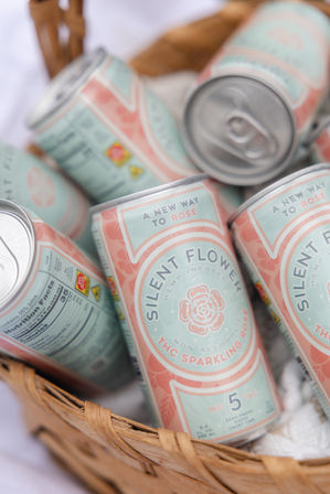 Pastel pink and mint cans of non-alcoholic THC sparkling rosé nestled in a wicker picnic basket, labels reading "A new way to rosé" — bright, summery canned rosé alternative.