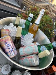 Metal ice bucket on a rooftop packed with ice and a cheerful mix of canned sparkling rosé, flavored sparkling water, and chilled wine and sparkling-water bottles with buildings and plants in the background
