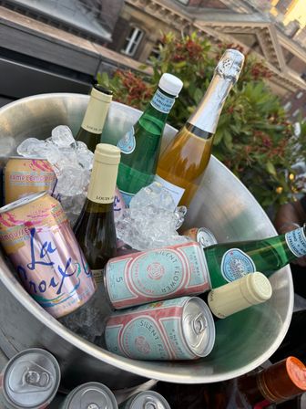 Metal ice bucket on a rooftop packed with ice and a cheerful mix of canned sparkling rosé, flavored sparkling water, and chilled wine and sparkling-water bottles with buildings and plants in the background