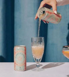 Hand pouring canned sparkling rosé into a crystal flute on a pastel tabletop with pink roses, cherries and a blue curtain backdrop.