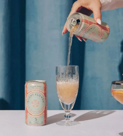 Hand pouring canned sparkling rosé into a crystal flute on a pastel tabletop with pink roses, cherries and a blue curtain backdrop.
