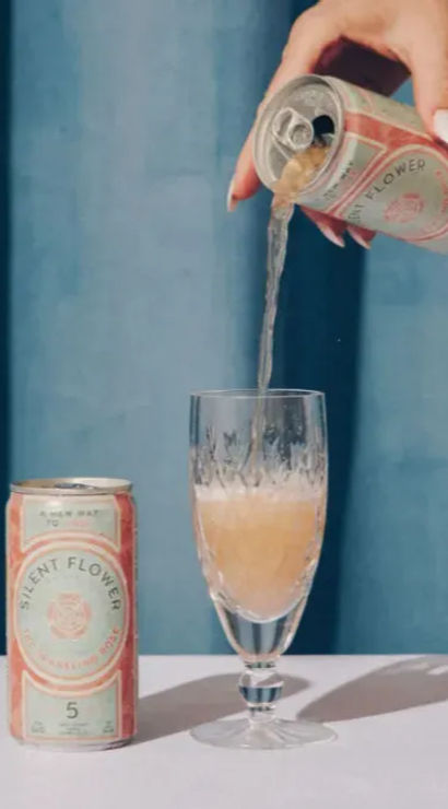 Hand pouring canned sparkling rosé into a crystal flute on a pastel tabletop with pink roses, cherries and a blue curtain backdrop.