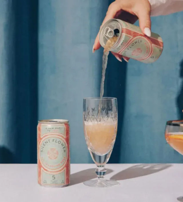 Hand pouring canned sparkling rosé into a crystal flute on a pastel tabletop with pink roses, cherries and a blue curtain backdrop.