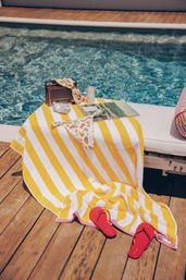 Poolside scene on a wooden deck with a yellow-and-white striped towel on a lounge, floral bikini pieces, red flip-flops, vintage radio, canned drink and magazine beside a turquoise pool.