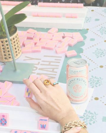 Hand wearing gold and turquoise rings and a braided bracelet reaches for pink domino-style game tiles on a pastel party table, beside a canned sparkling rosé and a small potted plant.