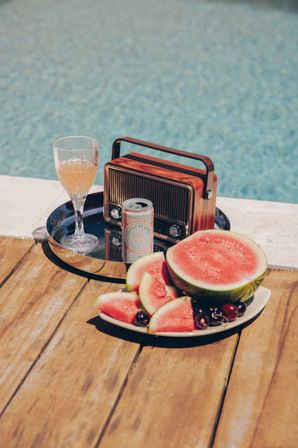 Sunny poolside summer scene on a wooden deck with a vintage portable radio and glass of juice on a metal tray next to a canned drink, and a plate of sliced watermelon and cherries