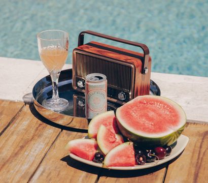 Sunny poolside summer scene on a wooden deck with a vintage portable radio and glass of juice on a metal tray next to a canned drink, and a plate of sliced watermelon and cherries