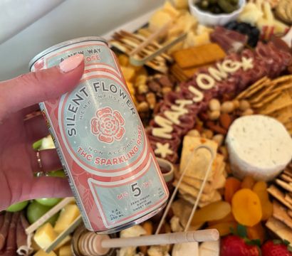 Hand with pale pink nails holding a pink can of THC sparkling rosé over a loaded charcuterie board with cheeses, crackers, olives, dried apricots and strawberries — colorful appetizer spread for a brunch or party