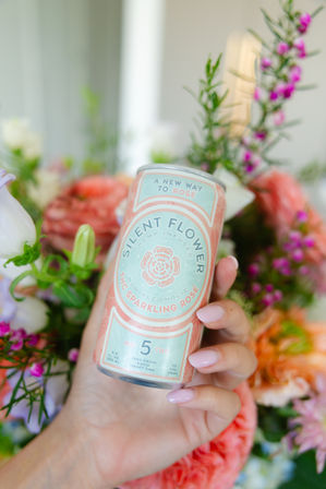 Close-up of a hand with pale pink nails holding a pastel pink-and-mint sparkling rosé can (hemp-infused), surrounded by a vibrant bouquet of pink and purple flowers — bright spring beverage shot.