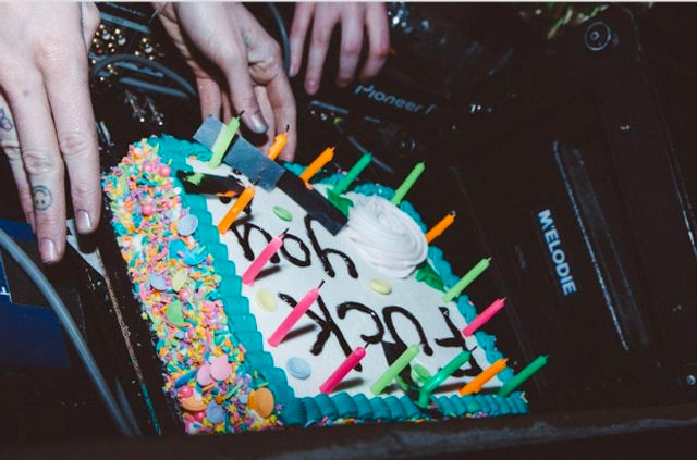 Colorful rectangular sprinkle cake with teal frosting border, multicolored candles and a handwritten message being placed on a DJ booth among hands and audio equipment